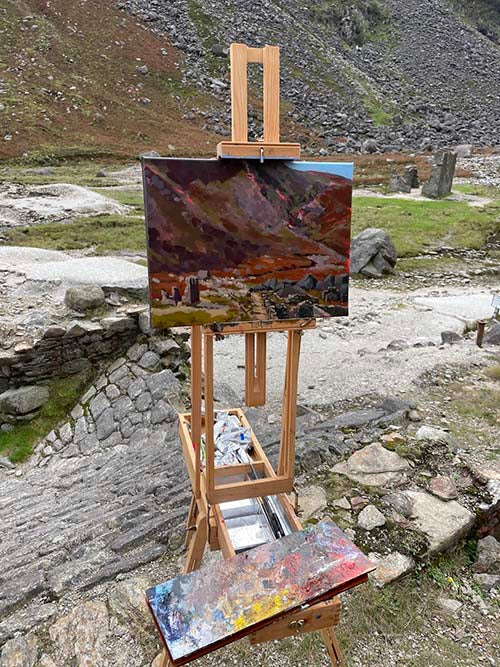 A painting is in progress and unfinished on an easel on location at the Miners village in Glendalough, the easel is situated on top of ruined structures in the background is a scree heavy valley slope.