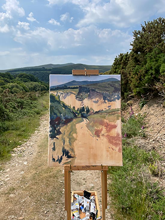 A painting is on an easel on a track through country side with mountains in the background. The painting has been started but is not finished.