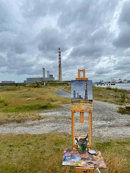 A painting on an easel outside in front of industrial chimneys in a waterfront scene in Dublin, the painting is not finished but is in progress.