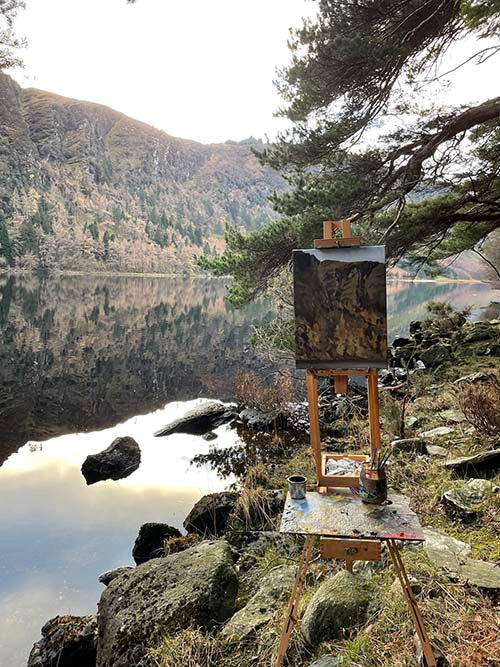 A painting is in progress on an easel at a lakes edge, the view is of the lake and cliff faces on the opposite side of the lake, location is the upper lake in Glendalough.