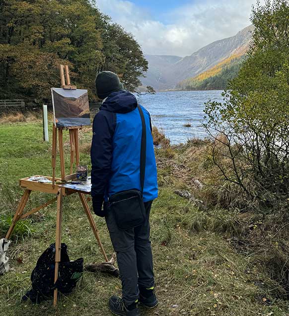 Artist Kieran Guckian painting at the shore of the upper lake in Glendalough. He is standing in front of an easel with a canvas on it.